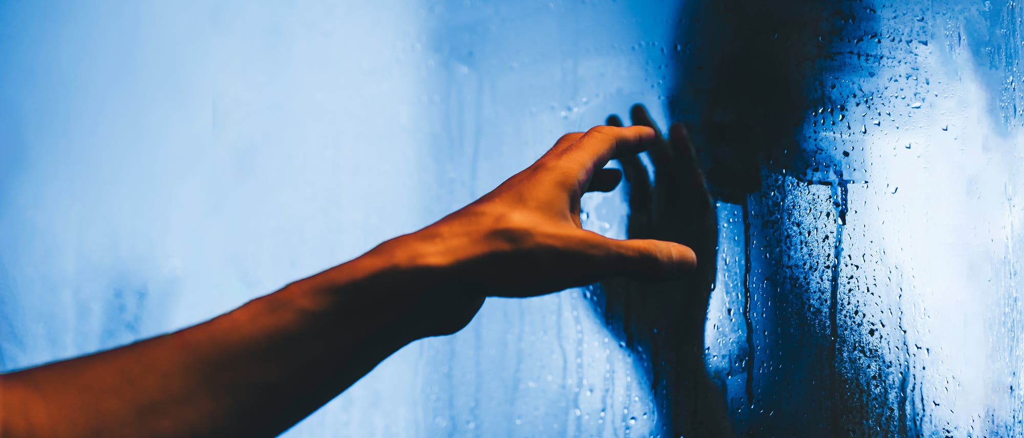 Eine Hand streckt sich in Richtung einer beschlagenen Glasscheibe, auf der sich Wassertropfen befinden. Im Hintergrund ist die verschwommene Silhouette einer Person zu erkennen. Die Szene ist in blauen Tönen gehalten. Eine Hand streckt sich in Richtung einer beschlagenen Glasscheibe, auf der sich Wassertropfen befinden. Im Hintergrund ist die verschwommene Silhouette einer Person zu erkennen. Die Szene ist in blauen Tönen gehalten.