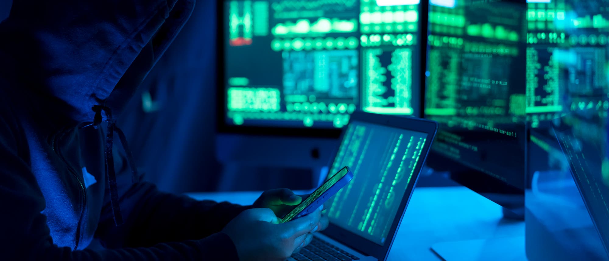 Eine Person in einem dunklen Raum sitzt vor mehreren Bildschirmen, die mit grünen Codezeilen gefüllt sind. Die Person trägt einen Kapuzenpullover und hält ein Smartphone in der Hand, während sie auf einen Laptop schaut. Die Szene ist in blauem Licht getaucht, was eine geheimnisvolle und technische Atmosphäre schafft. Themen wie Cyber-Sicherheit und Hacking werden angedeutet. Eine Person in einem dunklen Raum sitzt vor mehreren Bildschirmen, die mit grünen Codezeilen gefüllt sind. Die Person trägt einen Kapuzenpullover und hält ein Smartphone in der Hand, während sie auf einen Laptop schaut. Die Szene ist in blauem Licht getaucht, was eine geheimnisvolle und technische Atmosphäre schafft. Themen wie Cyber-Sicherheit und Hacking werden angedeutet.