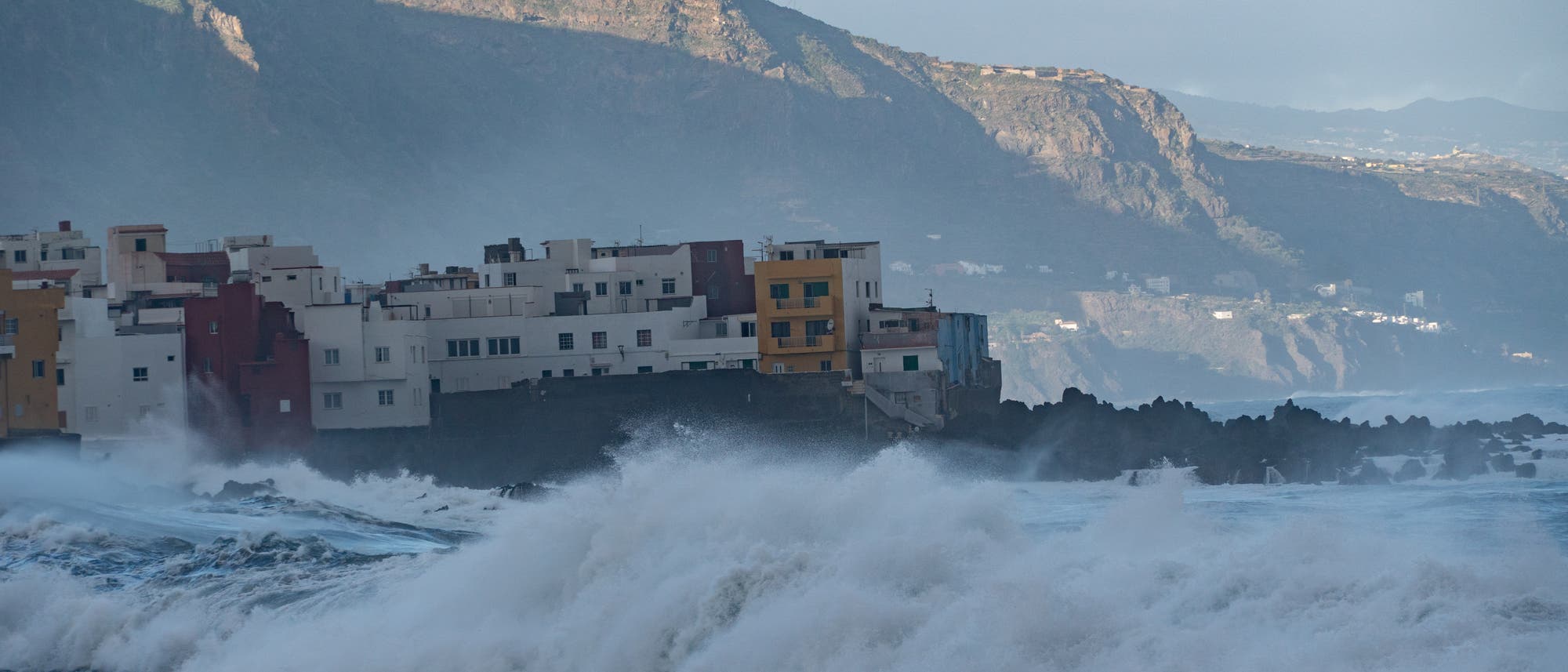 K&uuml;stenlandschaft mit st&uuml;rmischer See und hohen Wellen, die gegen eine Reihe bunter Geb&auml;ude schlagen. Im Hintergrund sind felsige Klippen zu sehen, die von der untergehenden Sonne beleuchtet werden. 