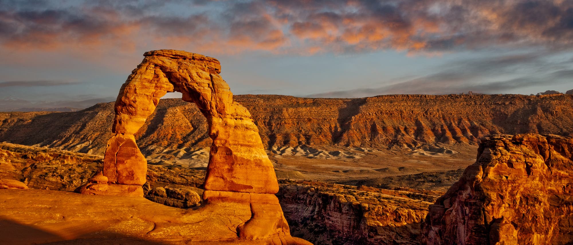 Eine beeindruckende Felsformation, bekannt als Delicate Arch, erhebt sich in der Wüstenlandschaft des Arches National Park in Utah, USA. Der Bogen aus rotem Sandstein wird von der warmen Abendsonne beleuchtet, die den Himmel in sanften Orange- und Rosatönen färbt. Im Hintergrund erstrecken sich zerklüftete Felsen und Hügel, die die Weite und Schönheit der Umgebung unterstreichen.