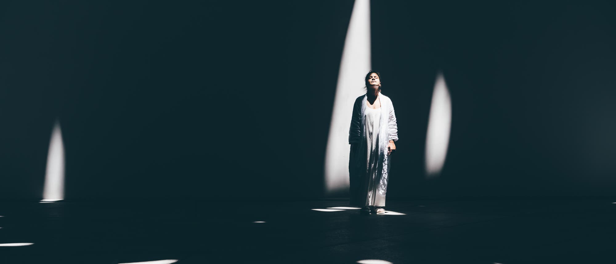 Eine Frau steht in einem großen, dunklen Raum, beleuchtet von schmalen Lichtstrahlen, die durch die Decke fallen. Sie trägt lange, helle Kleidung und hat den Kopf mit geschlossenen Augen leicht nach hinten gelegt, wie wenn sie das Licht genießt. Die Szene könnte Teil einer Theateraufführung sein.