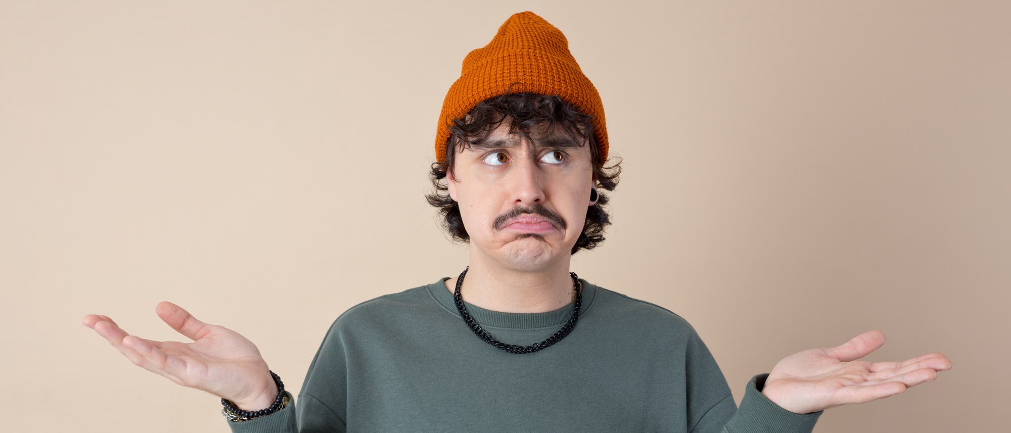 Eine Person mit einem orangefarbenen Strickm&uuml;tze und einem gr&uuml;nen Pullover hebt die Schultern und h&auml;lt die H&auml;nde seitlich hoch, als ob sie verwirrt oder unsicher ist. Der Hintergrund ist einfarbig beige.