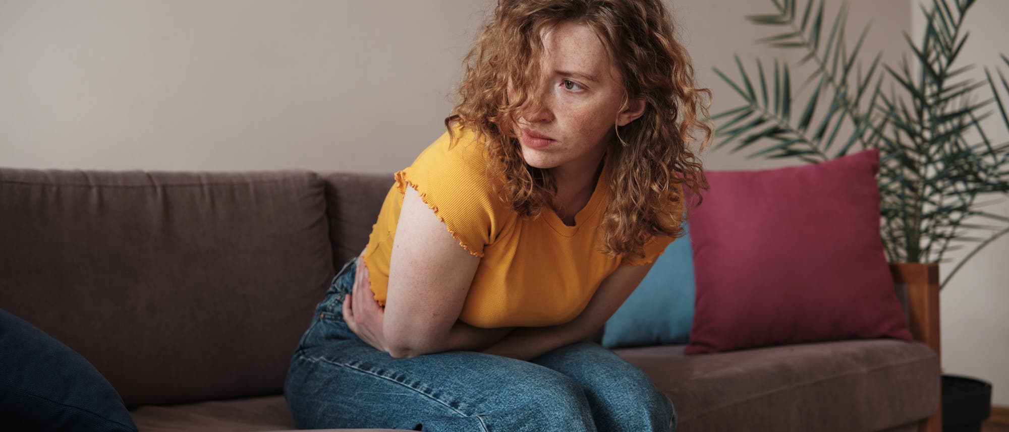 Eine Frau mit lockigem Haar sitzt auf einem Sofa und h&auml;lt sich den Bauch, als ob sie Schmerzen h&auml;tte. Sie tr&auml;gt ein gelbes T-Shirt und blaue Jeans. Im Hintergrund sind bunte Kissen und eine Zimmerpflanze zu sehen.