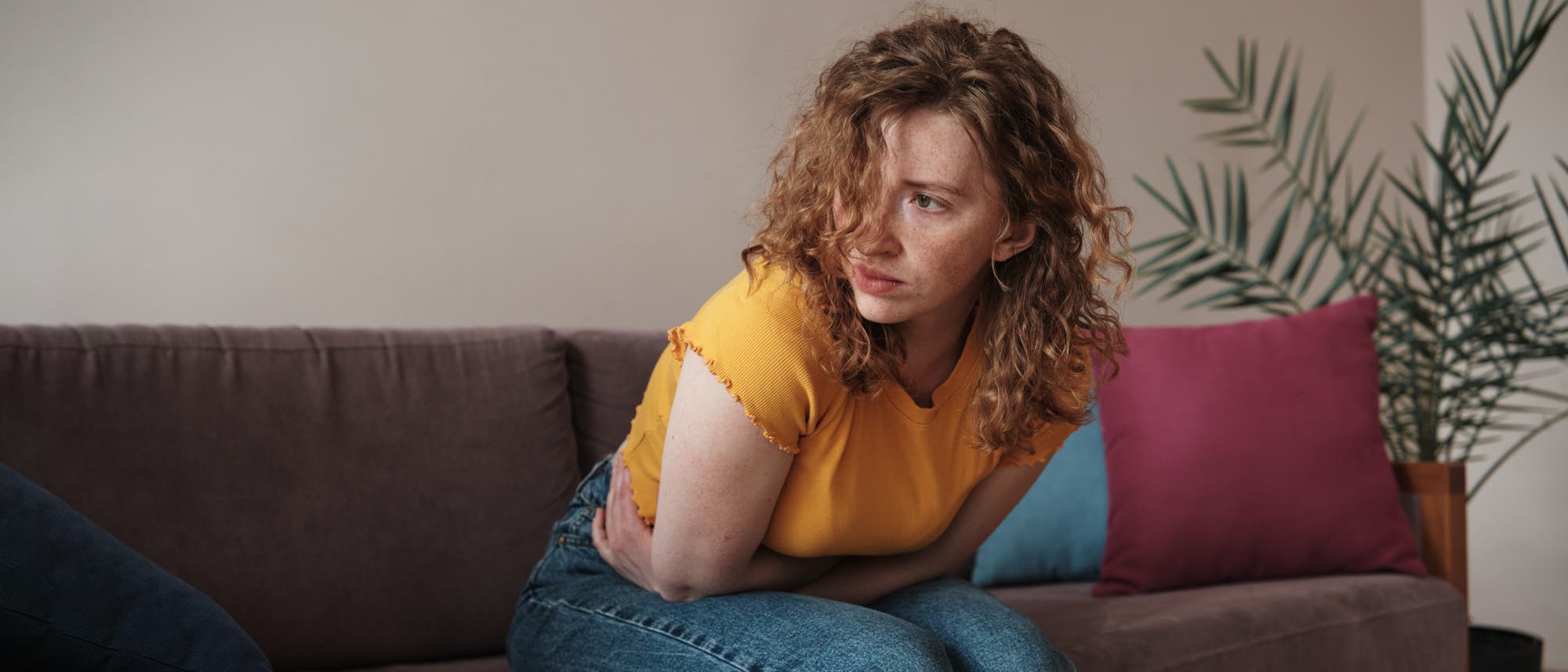 Eine Frau mit lockigem Haar sitzt auf einem Sofa und h&auml;lt sich den Bauch, als ob sie Schmerzen h&auml;tte. Sie tr&auml;gt ein gelbes T-Shirt und blaue Jeans. Im Hintergrund sind bunte Kissen und eine Zimmerpflanze zu sehen.