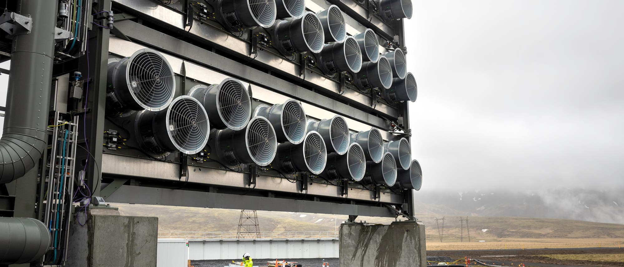 Eine industrielle Anlage zur CO2-Abscheidung in einer kargen Landschaft. Mehrere gro&szlig;e Ventilatoren sind in Reihen auf einer Metallstruktur montiert. Im Hintergrund sind neblige H&uuml;gel und ein bew&ouml;lkter Himmel zu sehen. Eine Person in Sicherheitskleidung geht auf dem Gel&auml;nde.