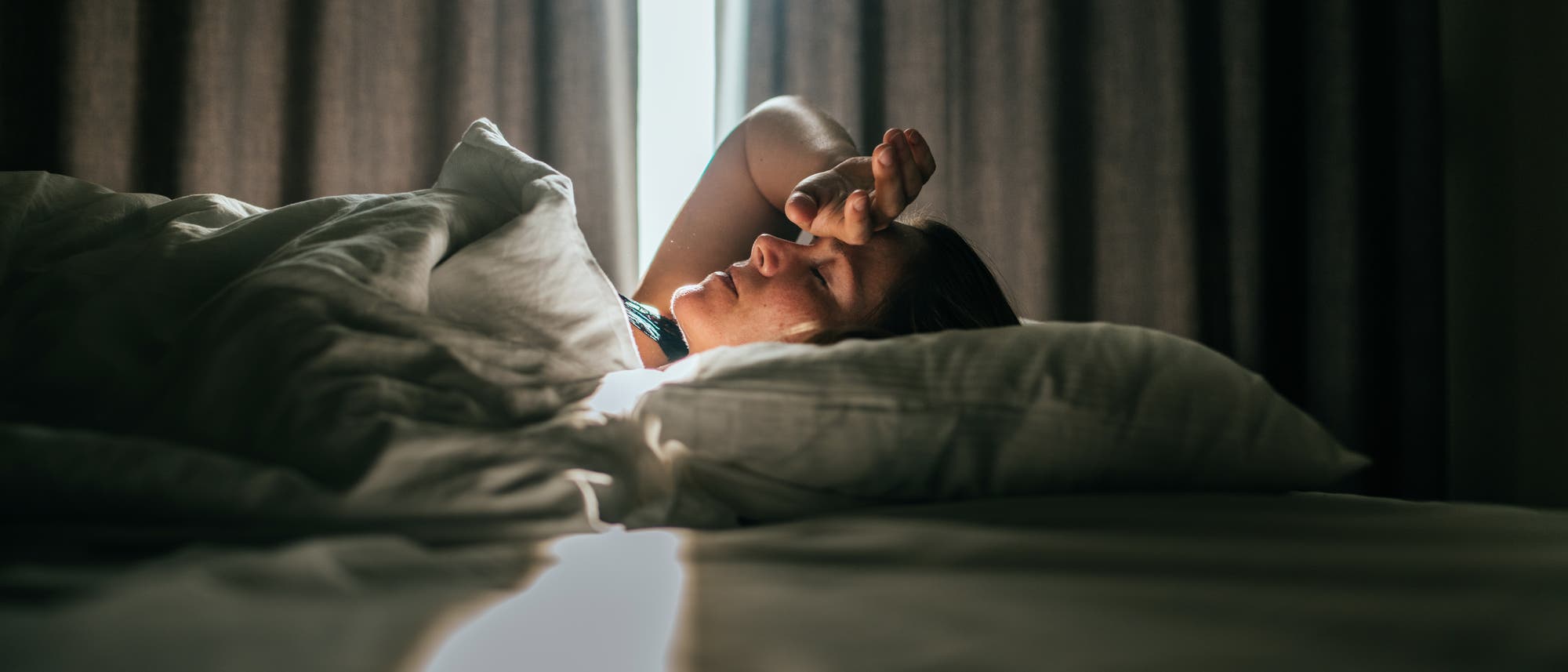 Eine Person liegt in einem Bett, teilweise von einer Decke bedeckt, mit einem Arm über der Stirn. Das Zimmer ist schwach beleuchtet, mit einem Lichtstrahl, der durch einen Spalt in den Vorhängen fällt und die Szene sanft erhellt. Die Atmosphäre wirkt ruhig und entspannt.