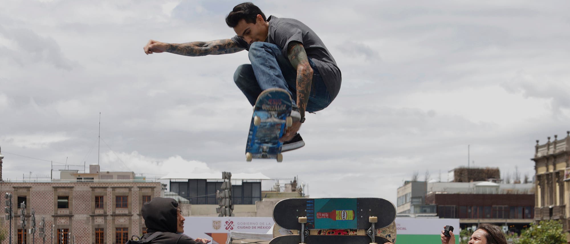 Ein Skateboarder f&uuml;hrt einen Trick in der Luft aus, w&auml;hrend er &uuml;ber eine Rampe springt. Im Hintergrund sind Geb&auml;ude und ein bew&ouml;lkter Himmel zu sehen. Zwei Personen beobachten die Szene, eine davon h&auml;lt eine Kamera.