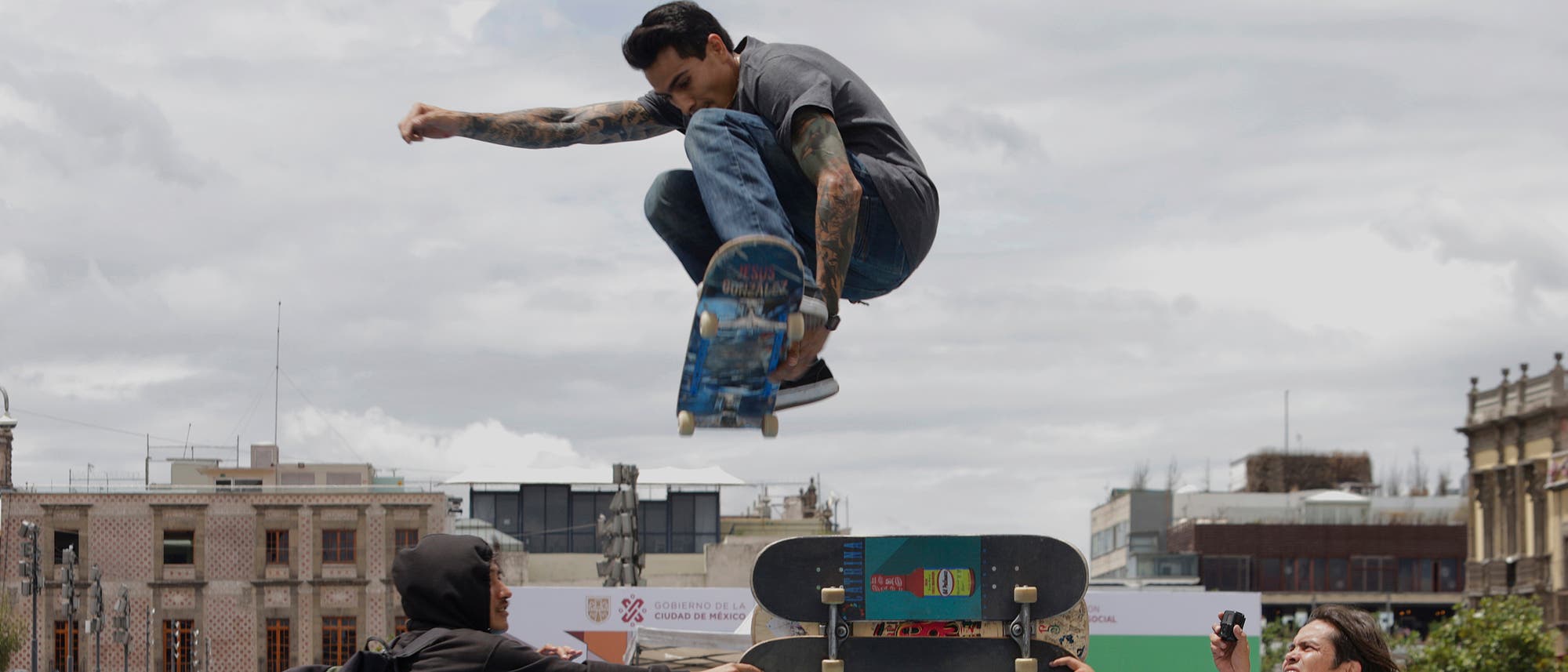 Ein Skateboarder führt einen Trick in der Luft aus, während er über eine Rampe springt. Im Hintergrund sind Gebäude und ein bewölkter Himmel zu sehen. Zwei Personen beobachten die Szene, eine davon hält eine Kamera. Ein Skateboarder führt einen Trick in der Luft aus, während er über eine Rampe springt. Im Hintergrund sind Gebäude und ein bewölkter Himmel zu sehen. Zwei Personen beobachten die Szene, eine davon hält eine Kamera.