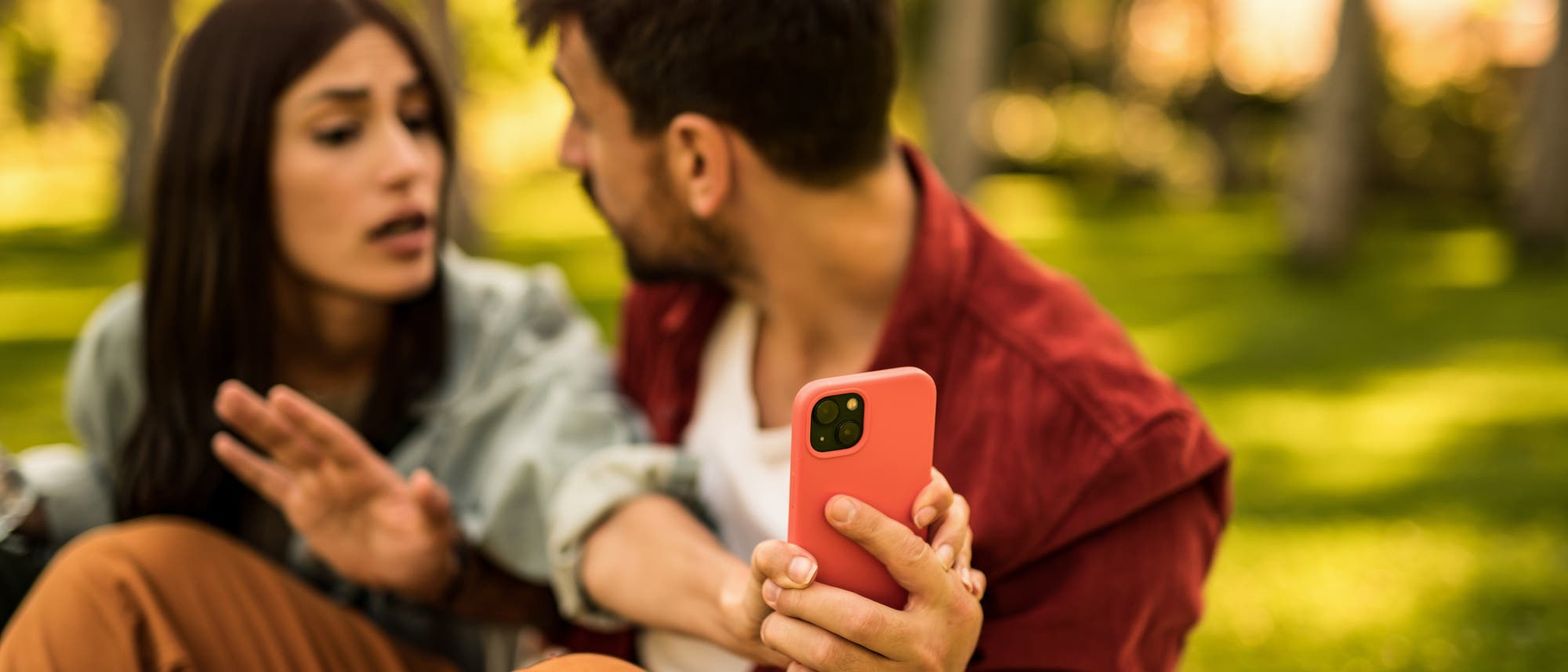 Ein junger Mann und eine junge Frau sitzen im Grünen, vermutlich auf einer Bank im Park. Die Frau greift nach einem Smartphone in der Hand des Mannes, aber er scheint es ihr nicht geben zu wollen. Die Stimmung zwischen den beiden wirkt angespannt.