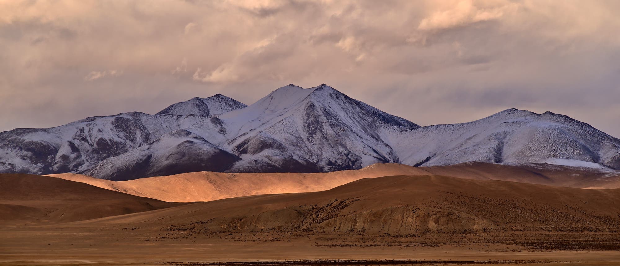 Eine weite, trockene Landschaft mit schneebedeckten Bergen im Hintergrund. Der Himmel ist teilweise bew&ouml;lkt, und die Sonne wirft ein warmes Licht auf die H&uuml;gel im Vordergrund. Die Szene vermittelt eine ruhige und majest&auml;tische Atmosph&auml;re.