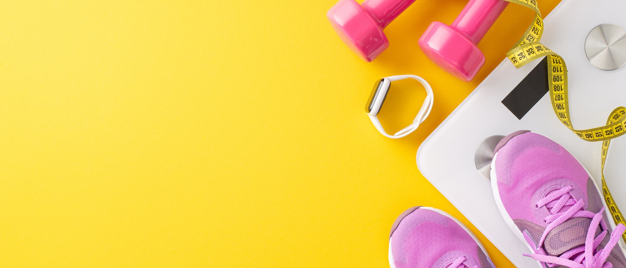 Ein Paar pinke Sportschuhe, zwei pinke Hanteln, ein Ma&szlig;band und eine Smartwatch liegen auf einer wei&szlig;en Waage vor einem gelben Hintergrund. Die Szene vermittelt ein Fitness- und Gesundheitsmotiv.