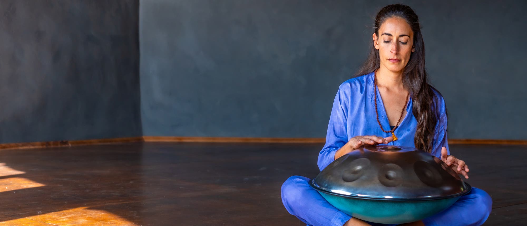 Eine Person sitzt in einem ruhigen Raum auf einem Teppich und spielt ein Handpan-Instrument. Sie trägt ein blaues Outfit und hat die Augen geschlossen, während sie das Instrument mit den Händen berührt. Der Raum ist spärlich beleuchtet, mit einem dunklen, strukturierten Hintergrund. Die Szene vermittelt eine Atmosphäre der Entspannung und Konzentration.