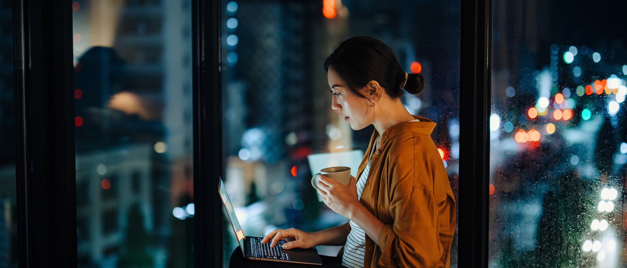 Eine Person sitzt nachts in einem Geb&auml;ude am Fenster und arbeitet an einem Laptop. Sie h&auml;lt eine Tasse in der Hand. Drau&szlig;en sind verschwommene Lichter einer Stadt zu sehen, die eine lebendige n&auml;chtliche Atmosph&auml;re schaffen.