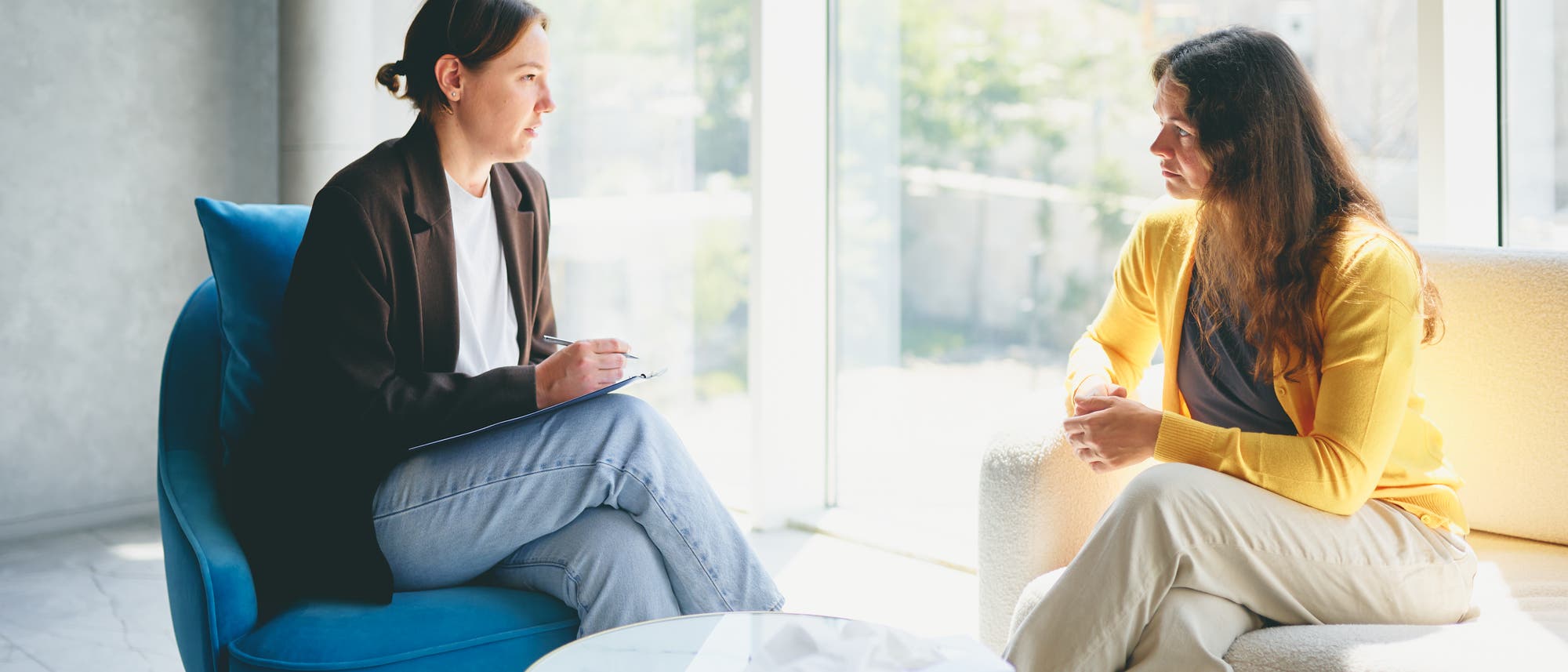 Zwei Personen sitzen in einem hellen Raum mit großen Fenstern. Eine Person hält ein Klemmbrett und scheint Notizen zu machen, während sie der anderen Person zuhört. Die zweite Person sitzt entspannt auf einem Sofa. Auf dem Tisch zwischen ihnen liegt eine Box mit Taschentüchern. Die Szene wirkt ruhig und professionell.