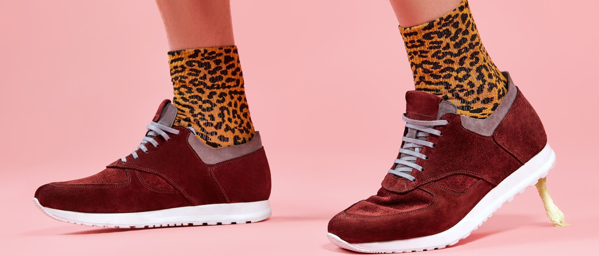 Eine Person trägt rote Wildleder-Sneaker mit weißen Sohlen und grauen Schnürsenkeln. Darin stecken gelb-schwarz gemusterten Socken mit Leopardenmuster. Der Hintergrund ist einfarbig rosa. Der hintere Schuh hat einen Kaugummi an der Sohle kleben.