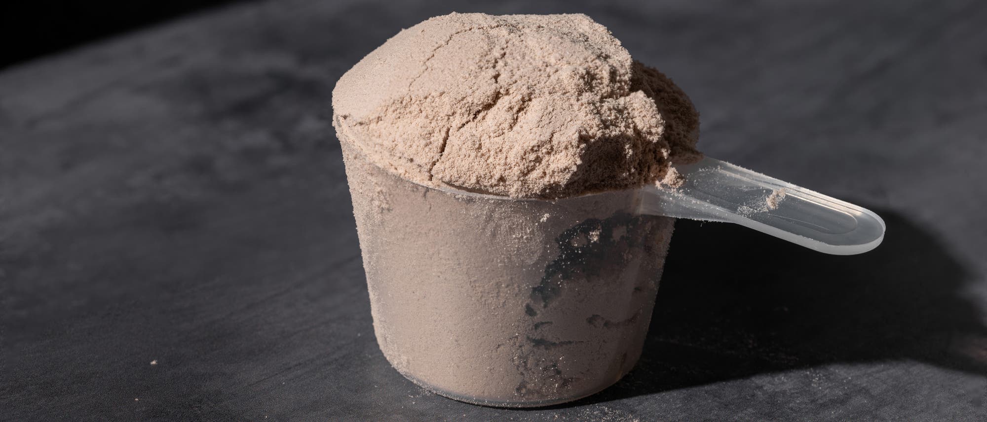 Ein transparenter Messlöffel, gefüllt mit beigem Pulver, liegt auf einer dunklen Oberfläche. Das Pulver ist bis zum Rand des Löffels gehäuft. Die Szene ist gleichmäßig ausgeleuchtet, wodurch die Textur des Pulvers deutlich sichtbar wird.