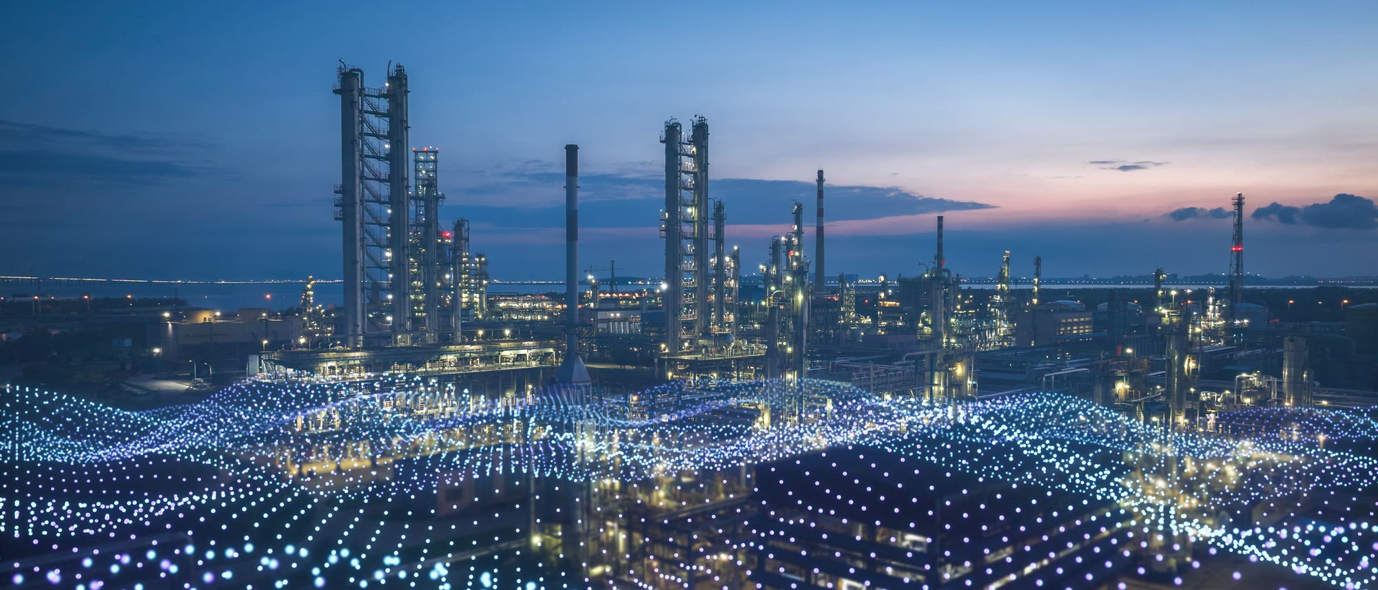 Industrieanlage bei Dämmerung mit mehreren hohen Türmen und Schornsteinen, die sich gegen den Abendhimmel abheben. Im Vordergrund sind leuchtende, wellenförmige Muster zu sehen, die möglicherweise digitale Datenströme symbolisieren. Die Szene vermittelt eine Verbindung von industrieller Infrastruktur und moderner Technologie.