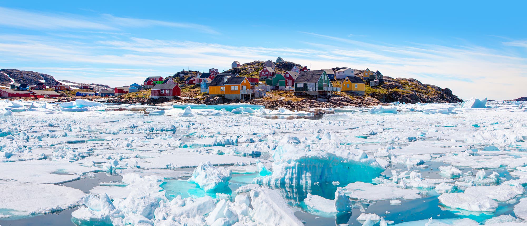 Bunte Häuser auf einem felsigen Hügel in einem grönländischen Dorf, umgeben von einer weiten, eisbedeckten Landschaft. Im Vordergrund sind große Eisblöcke und schimmerndes blaues Wasser zu sehen. Der Himmel ist klar und blau, was die kühle, ruhige Atmosphäre der arktischen Umgebung unterstreicht.