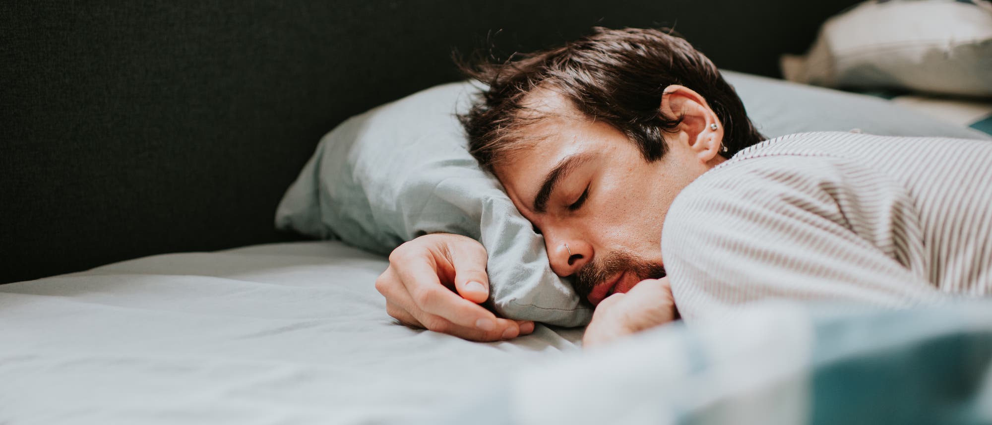 Eine Person liegt schlafend auf der Seite in einem Bett mit hellblauer Bettwäsche. Der Kopf ruht auf einem Kissen, und die Person trägt ein gestreiftes Oberteil. Im Hintergrund ist ein Teil des Bettrahmens und ein weiteres Kissen zu sehen. Eine Person liegt schlafend auf der Seite in einem Bett mit hellblauer Bettwäsche. Der Kopf ruht auf einem Kissen, und die Person trägt ein gestreiftes Oberteil. Im Hintergrund ist ein Teil des Bettrahmens und ein weiteres Kissen zu sehen.