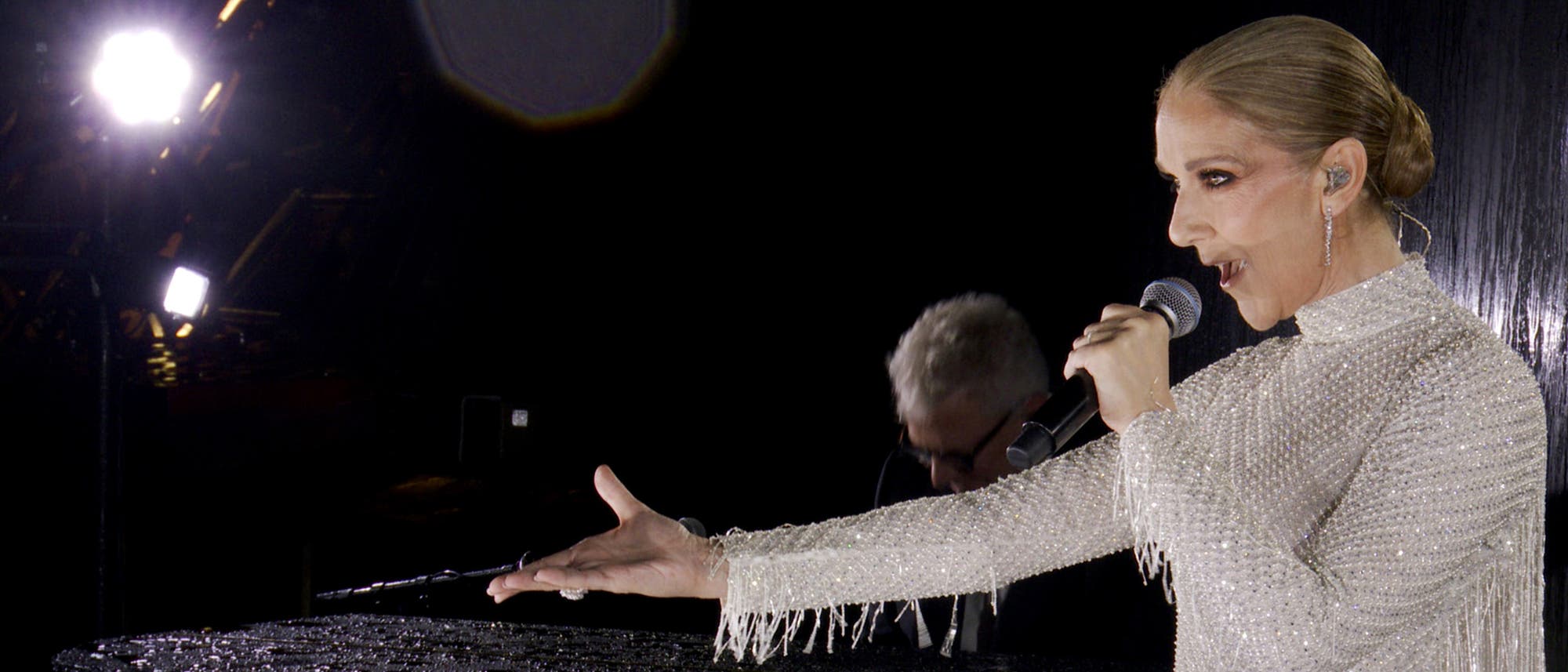 Celine Dion in einem glitzernden, weißen Kleid steht auf dem Eiffelturm und singt in ein Mikrofon. Ihre rechte Hand ist ausgestreckt, während sie performt. Im Hintergrund ist ein Pianist zu sehen, der auf einem schwarzen Flügel spielt. Die Umgebung erscheint dunkel, mit einigen hellen Lichtern im Hintergrund.
Celine Dion in einem glitzernden, weißen Kleid steht auf dem Eiffelturm und singt in ein Mikrofon. Ihre rechte Hand ist ausgestreckt, während sie performt. Im Hintergrund ist ein Pianist zu sehen, der auf einem schwarzen Flügel spielt. Die Umgebung erscheint dunkel, mit einigen hellen Lichtern im Hintergrund.