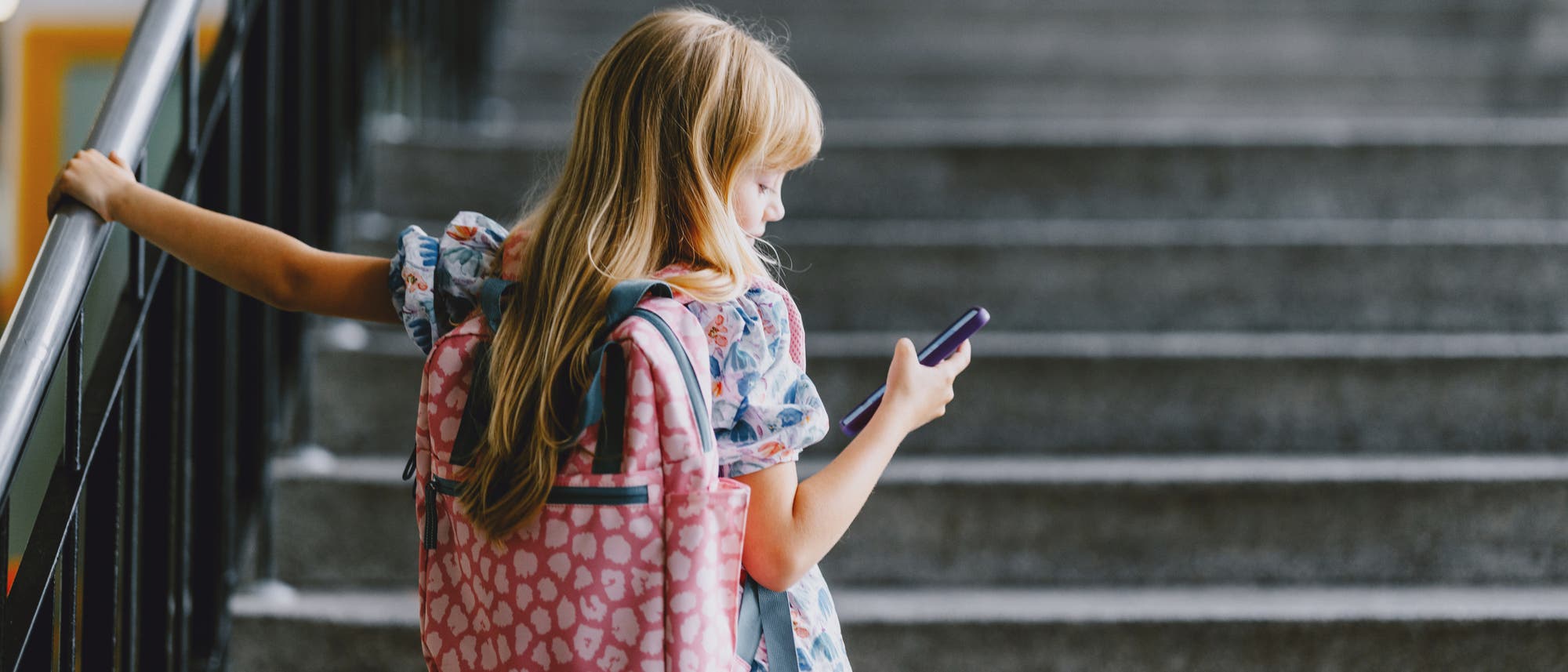 Ein Kind mit einem rosa Rucksack steht auf einer Treppe und schaut auf ein Smartphone. Es hält sich mit einer Hand am Geländer fest. Die Umgebung wirkt wie ein Schulgebäude.