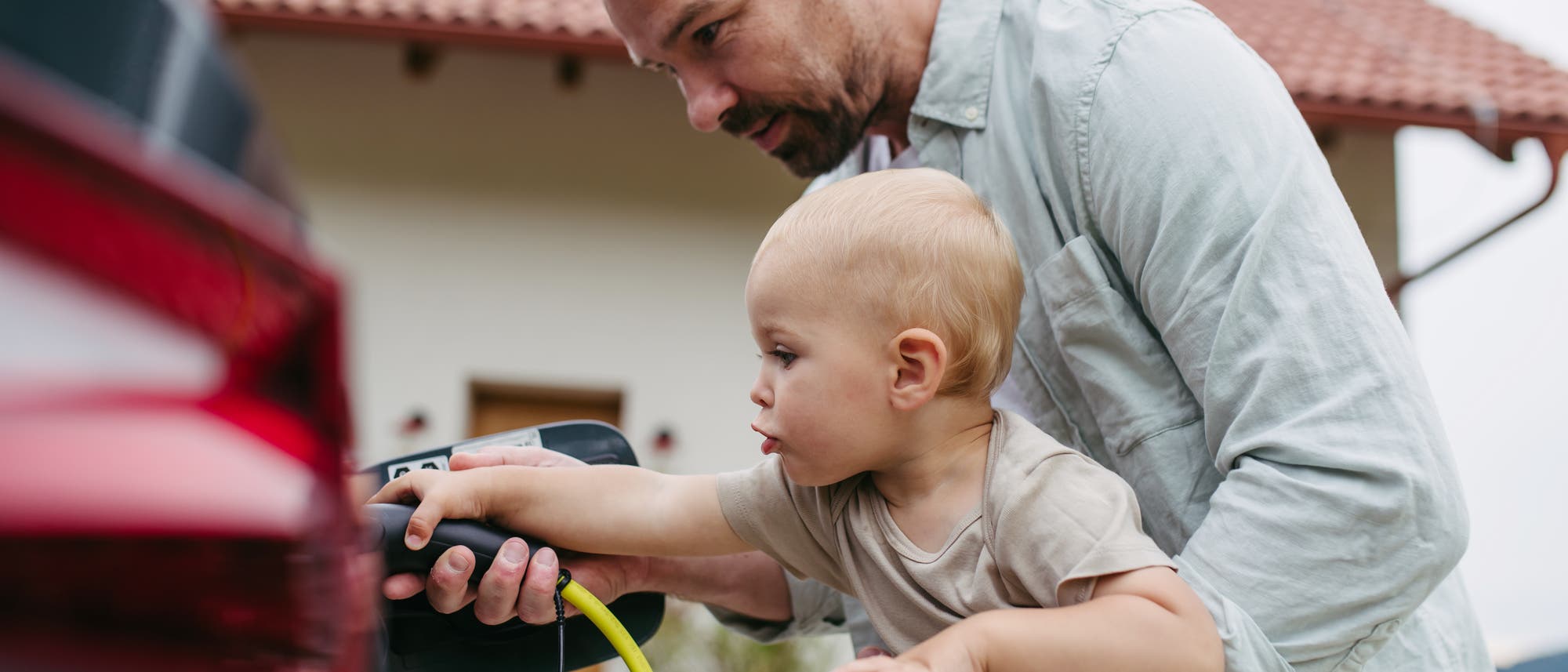 Ein Erwachsener h&auml;lt ein Kleinkind, w&auml;hrend sie gemeinsam ein Elektroauto aufladen. Der Erwachsene zeigt dem Kind, wie der Ladestecker in das Auto gesteckt wird. Im Hintergrund ist ein Haus mit einem roten Ziegeldach zu sehen. Die Szene vermittelt ein Gef&uuml;hl von Lernen und Zusammenarbeit.