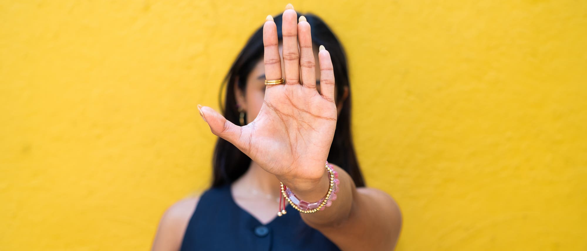 Eine Person steht vor einer gelben Wand und hebt die Handfläche zur Kamera, als ob sie ein Stoppsignal gibt. Die Person trägt ein dunkelblaues ärmelloses Oberteil und mehrere bunte Armbänder. Der Fokus liegt auf der Hand, die den Großteil des Bildes einnimmt. Eine Person steht vor einer gelben Wand und hebt die Handfläche zur Kamera, als ob sie ein Stoppsignal gibt. Die Person trägt ein dunkelblaues ärmelloses Oberteil und mehrere bunte Armbänder. Der Fokus liegt auf der Hand, die den Großteil des Bildes einnimmt.