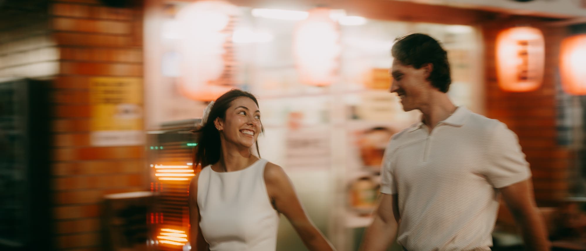 Ein l&auml;chelndes Paar geht Hand in Hand durch eine belebte Stra&szlig;e bei Nacht. Im Hintergrund sind verschwommene Lichter und Schilder eines Restaurants zu sehen, die eine lebendige und fr&ouml;hliche Atmosph&auml;re schaffen.