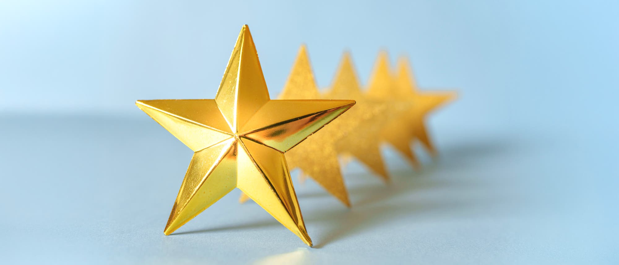 Eine Reihe von f&uuml;nf goldenen, dreidimensionalen Sternen, die in einer diagonalen Linie angeordnet sind. Der vorderste Stern ist scharf fokussiert, w&auml;hrend die anderen Sterne im Hintergrund unscharf sind. Die Sterne haben eine gl&auml;nzende Oberfl&auml;che und sind auf einem hellblauen Hintergrund platziert.