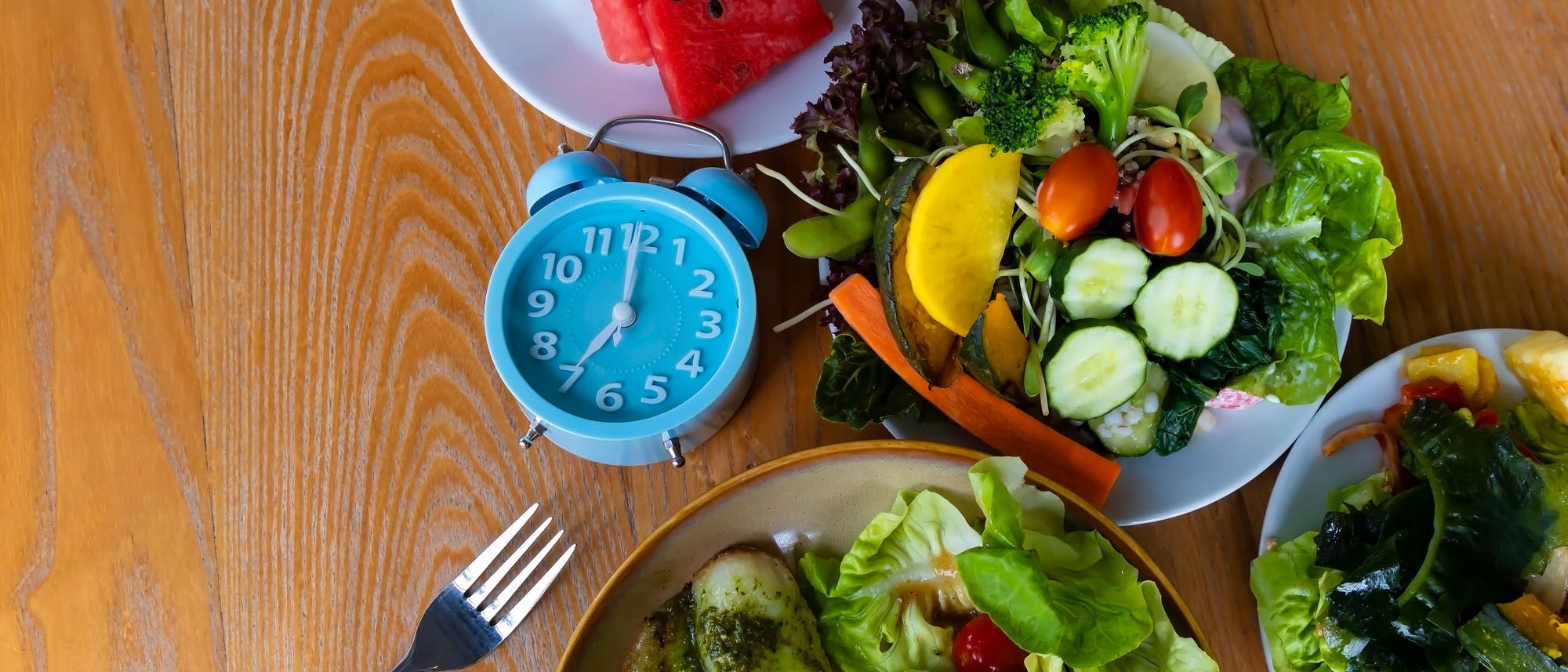 Ein Holztisch mit einer Auswahl an frischen Lebensmitteln: Ein Teller mit gemischtem Salat, Gurken, Tomaten und Karotten; ein weiterer Teller mit Wassermelonen- und Drachenfruchtstücken; ein Teller mit Fischfilet, Pommes frites und Dip-Sauce. Eine blaue Uhr zeigt 7:00 Uhr an. Links liegt eine Gabel.