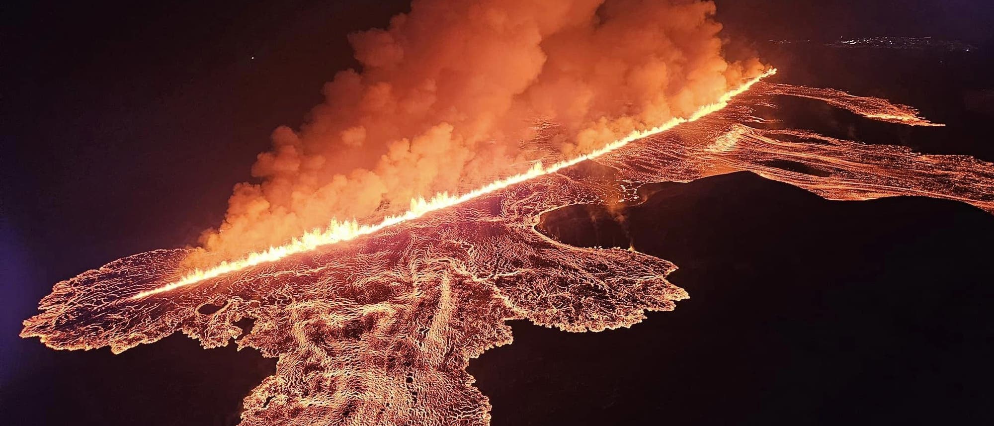 Luftaufnahme eines Vulkanausbruchs bei Nacht. Eine lange, glühende Lavafront zieht sich diagonal durch das Bild, umgeben von dichten Rauchwolken, die in den Nachthimmel aufsteigen. Die Lava leuchtet intensiv orange und rot, während sie sich über die dunkle Landschaft ausbreitet. Im Hintergrund sind schwach beleuchtete Stadtlichter zu erkennen.