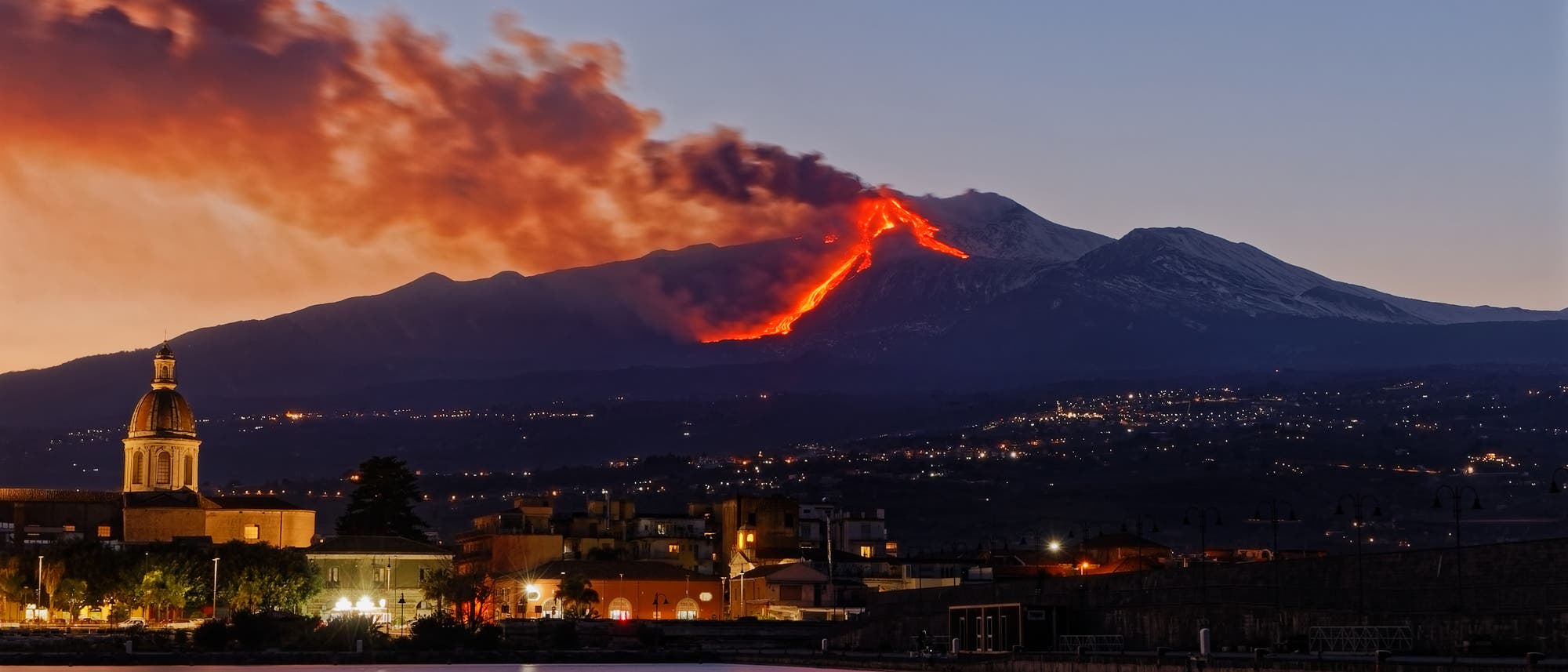 Eine n&auml;chtliche Landschaft zeigt einen Vulkan, der Lava ausst&ouml;&szlig;t, w&auml;hrend Rauch in den Himmel aufsteigt. Im Vordergrund ist eine Stadt mit beleuchteten Geb&auml;uden und einer Kirche mit Kuppel zu sehen. Der Vulkan leuchtet rot-orange, was einen starken Kontrast zum dunklen Himmel und der ruhigen Wasseroberfl&auml;che im Vordergrund bildet.