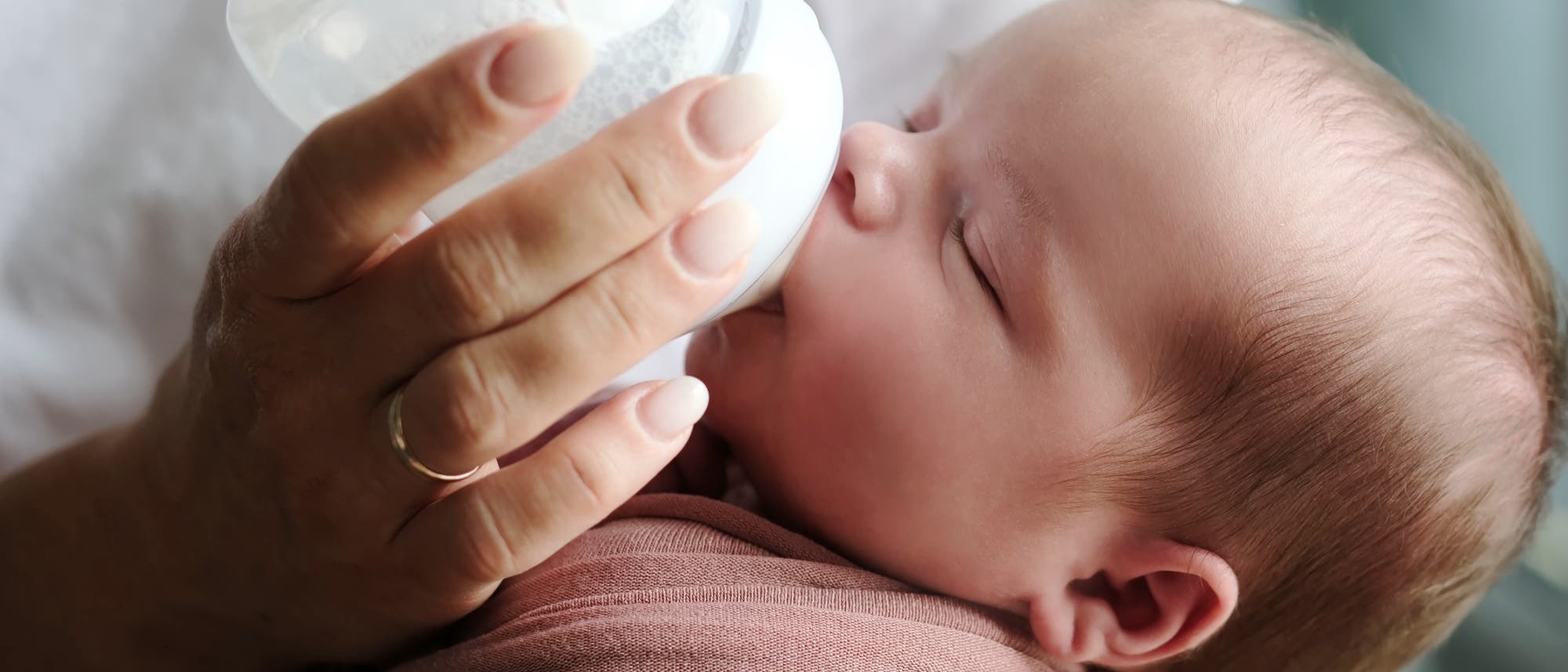 Eine Person h&auml;lt ein Baby, das aus einer Flasche trinkt. Das Baby ist in ein rosafarbenes Tuch gewickelt und hat die Augen geschlossen. Die Szene vermittelt Ruhe und F&uuml;rsorge.