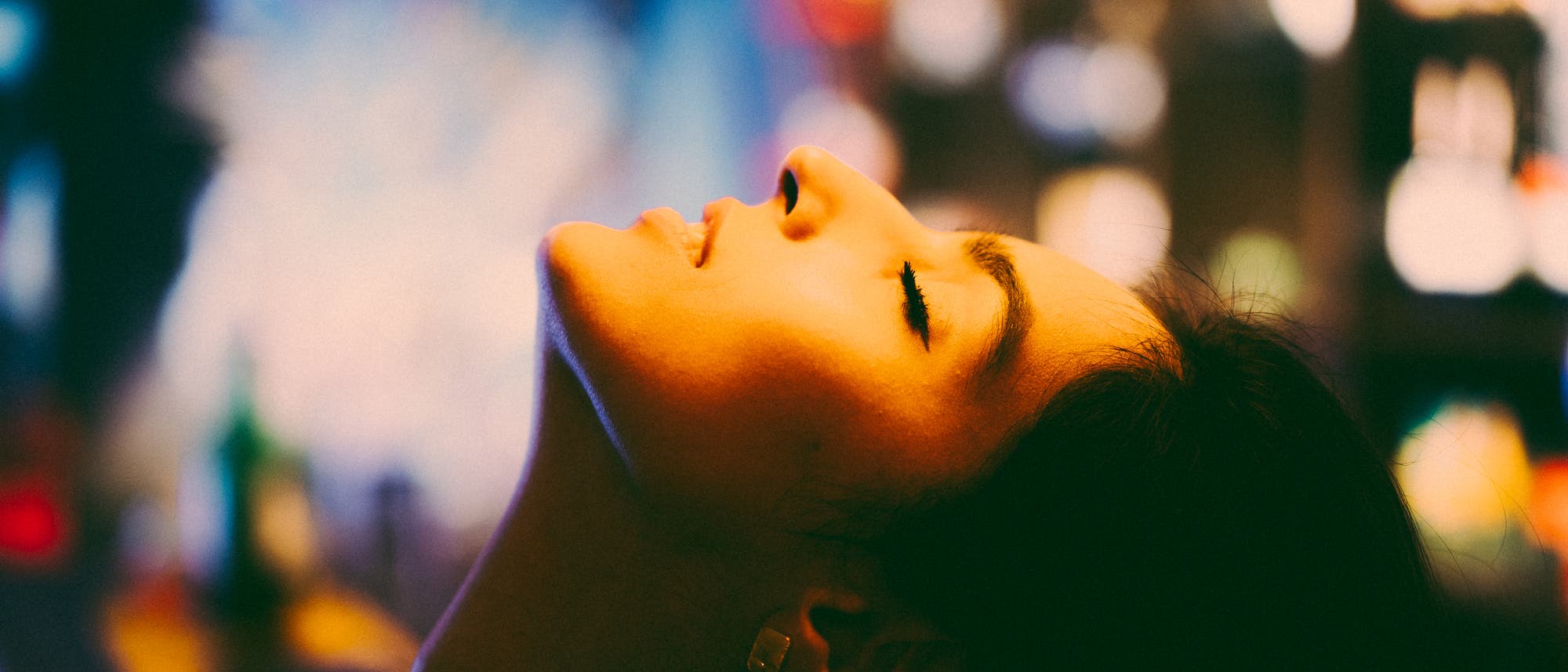 Eine Person mit geschlossenen Augen neigt den Kopf nach hinten, umgeben von einem unscharfen, farbenfrohen Hintergrund. Die Szene vermittelt ein Gef&uuml;hl von Entspannung und Gelassenheit. Die Beleuchtung erzeugt warme, weiche Schatten auf dem Gesicht.
