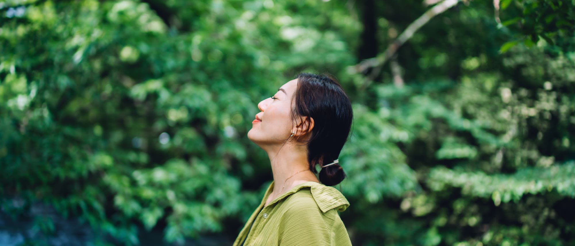 Eine Person steht in einem gr&uuml;nen Wald und genie&szlig;t die Natur. Sie tr&auml;gt eine gr&uuml;ne Jacke und hat die Augen geschlossen, w&auml;hrend sie tief einatmet. Im Hintergrund sind dichte B&auml;ume und ein flie&szlig;ender Bach zu sehen. Die Szene vermittelt Ruhe und Entspannung in einer nat&uuml;rlichen Umgebung.