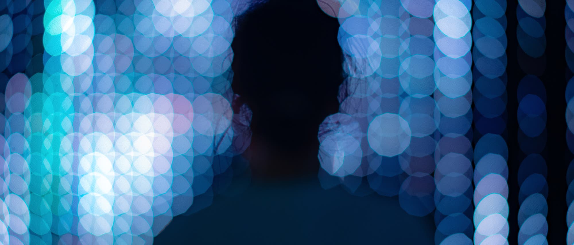 Eine unscharfe Silhouette einer Person steht vor einem Hintergrund aus verschwommenen, leuchtenden blauen und weißen Lichtern. Die Lichter sind in einem Bokeh-Effekt dargestellt, was eine traumhafte und abstrakte Atmosphäre erzeugt. Eine unscharfe Silhouette einer Person steht vor einem Hintergrund aus verschwommenen, leuchtenden blauen und weißen Lichtern. Die Lichter sind in einem Bokeh-Effekt dargestellt, was eine traumhafte und abstrakte Atmosphäre erzeugt.