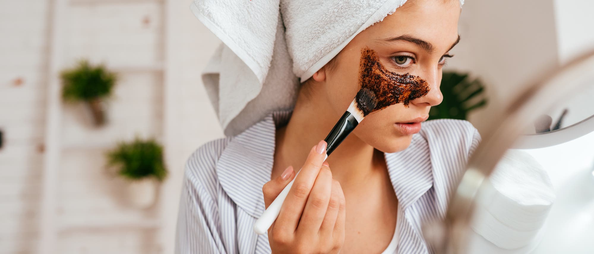 Eine M&auml;dchen m&ouml;chte ihre Haut pflegen und reinigen und tr&auml;gt mit einem Pinsel eine Gesichtsmaske auf die Wange auf. Bei der Skincareroutine f&uuml;r reine Haut hat sie ein Handtuch um den Kopf gewickelt und steht vor einem Spiegel. 