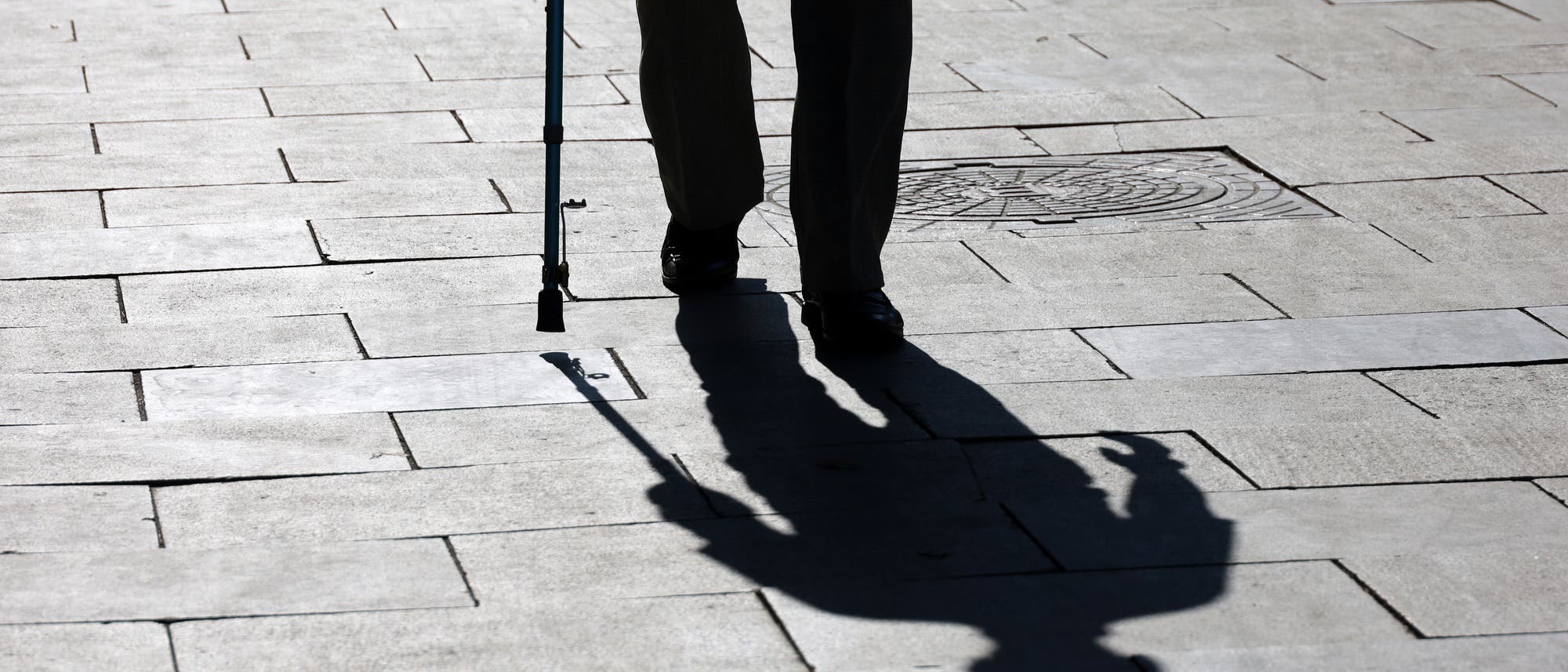 Eine Person geht mit einem Gehstock über einen gepflasterten Bürgersteig. Die tiefstehende Sonne wirft einen langen Schatten der Person auf den Boden. Eine Person geht mit einem Gehstock über einen gepflasterten Bürgersteig. Die tiefstehende Sonne wirft einen langen Schatten der Person auf den Boden.