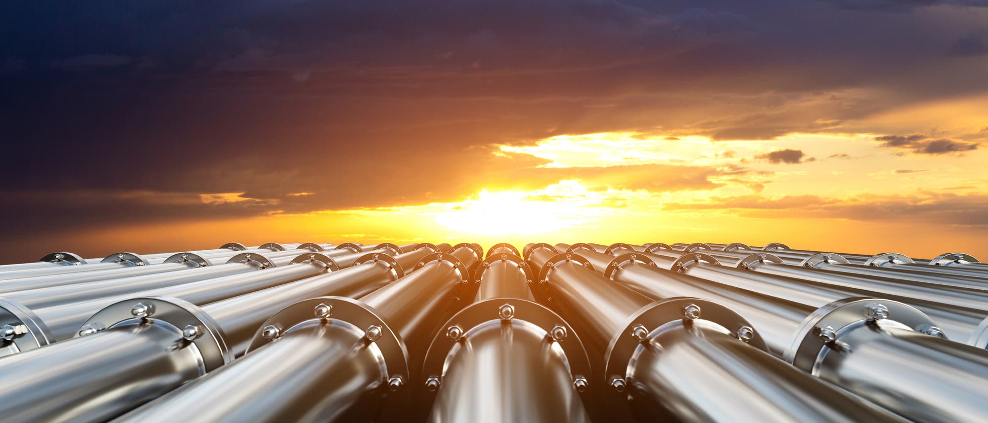 Eine Reihe von gl&auml;nzenden Metallrohren erstreckt sich in die Ferne, w&auml;hrend im Hintergrund ein dramatischer Sonnenuntergang den Himmel in warmen Gelb- und Oranget&ouml;nen f&auml;rbt. Die Rohre reflektieren das Licht und erzeugen einen Kontrast zu den dunklen Wolken am Himmel.