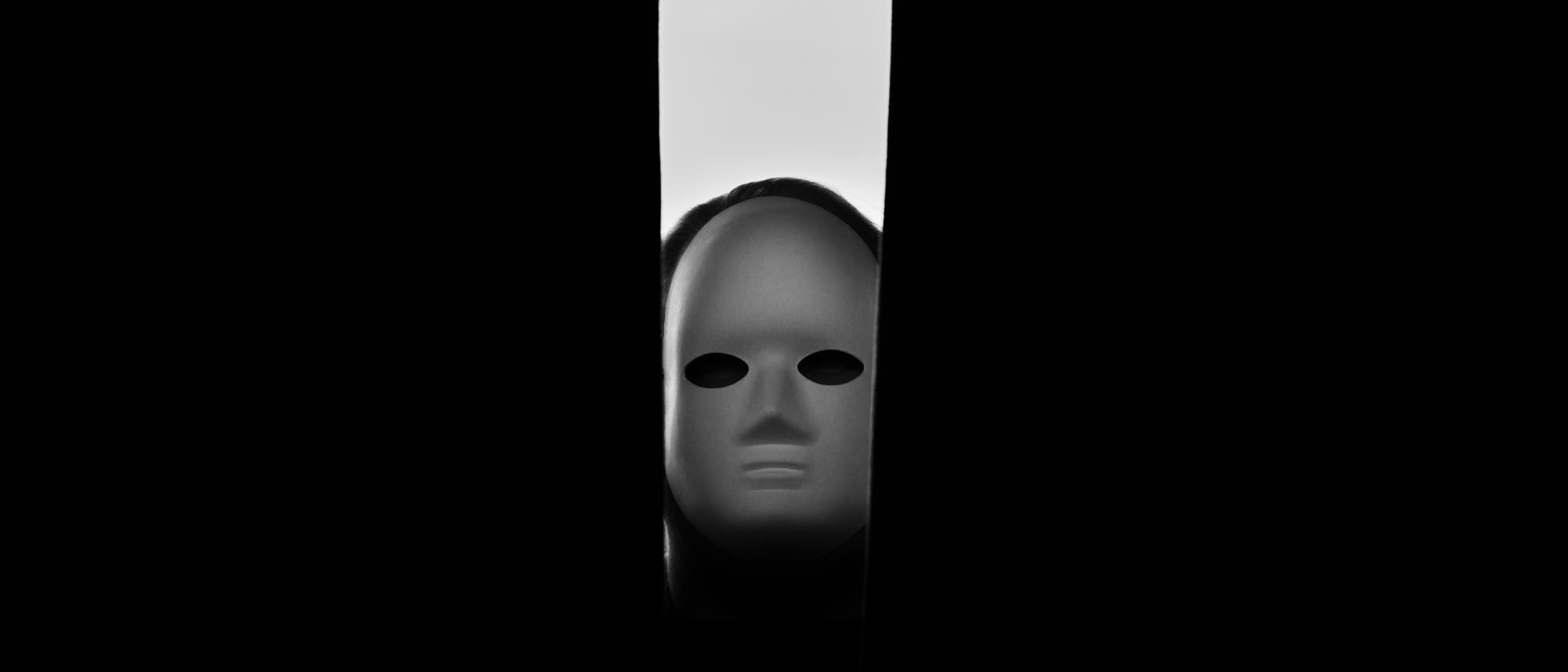 Eine wei&szlig;e Maske mit leeren Augen&ouml;ffnungen schaut durch einen schmalen Spalt zwischen zwei dunklen Vorh&auml;ngen. Die Szene ist in Schwarz-Wei&szlig; gehalten und erzeugt eine geheimnisvolle und leicht unheimliche Atmosph&auml;re.