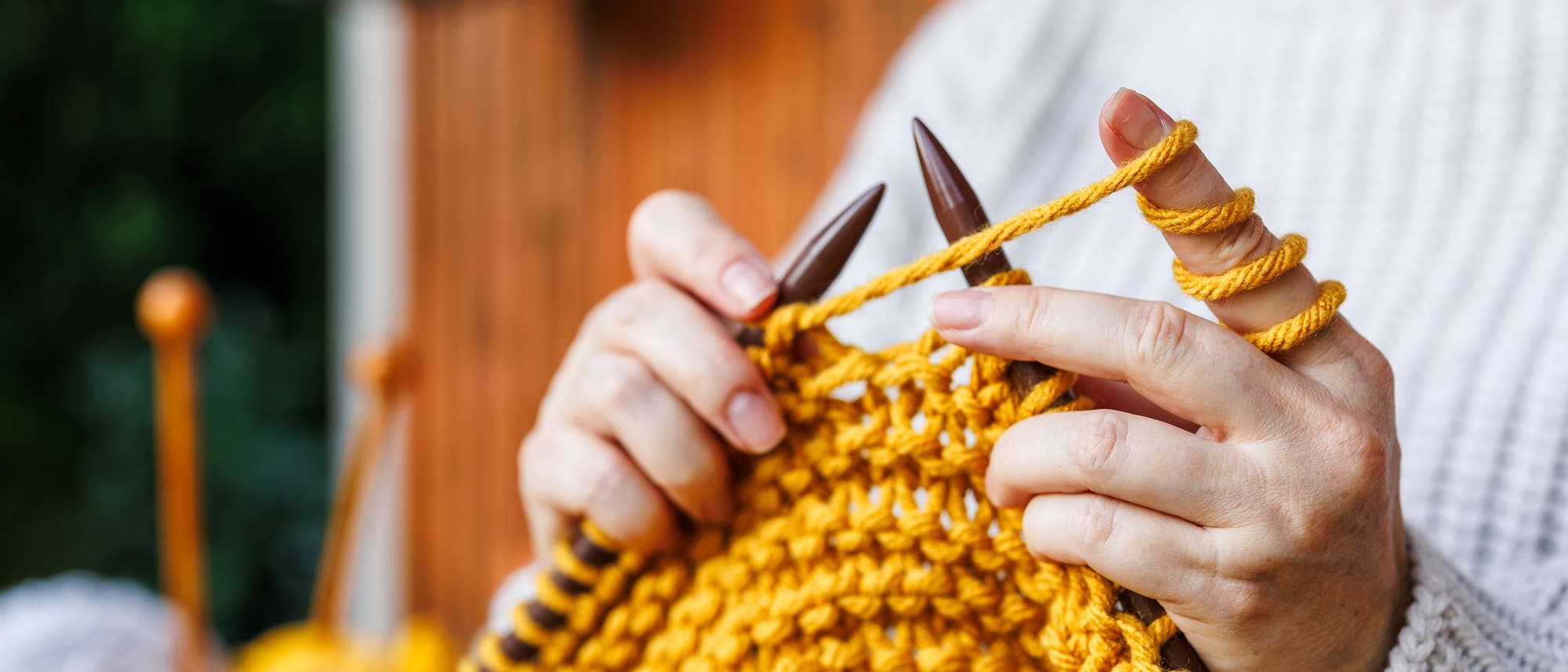 Eine Person strickt mit gelber Wolle und Holzstricknadeln. Die Hände sind im Fokus, während der Hintergrund unscharf ist. Die Person trägt einen cremefarbenen Pullover. Im Hintergrund sind weitere Wollknäuel zu sehen. Eine Person strickt mit gelber Wolle und Holzstricknadeln. Die Hände sind im Fokus, während der Hintergrund unscharf ist. Die Person trägt einen cremefarbenen Pullover. Im Hintergrund sind weitere Wollknäuel zu sehen.