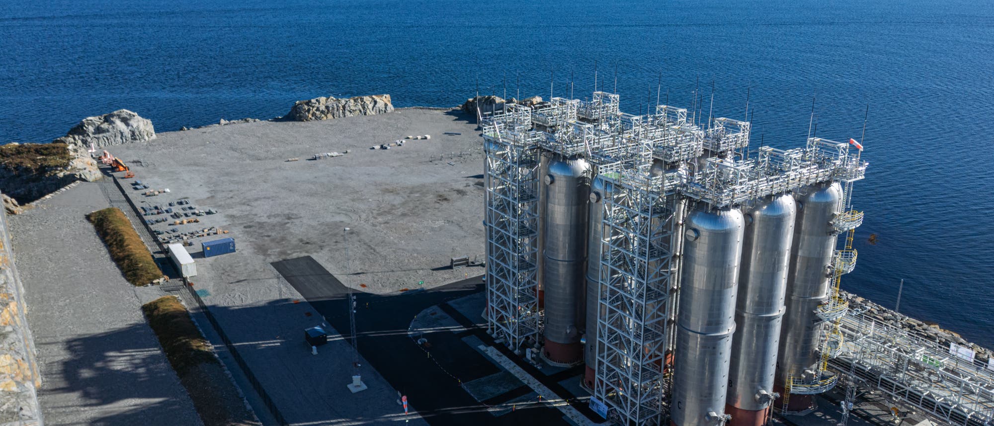 Industrieanlage an der Küste mit mehreren großen, silbernen Tanks und Rohrleitungen. Die Anlage befindet sich auf einem weitläufigen, gepflasterten Gelände neben einem tiefblauen Gewässer. Im Hintergrund sind Hügel und eine bewaldete Landschaft zu sehen. Der Himmel ist klar mit wenigen Wolken.