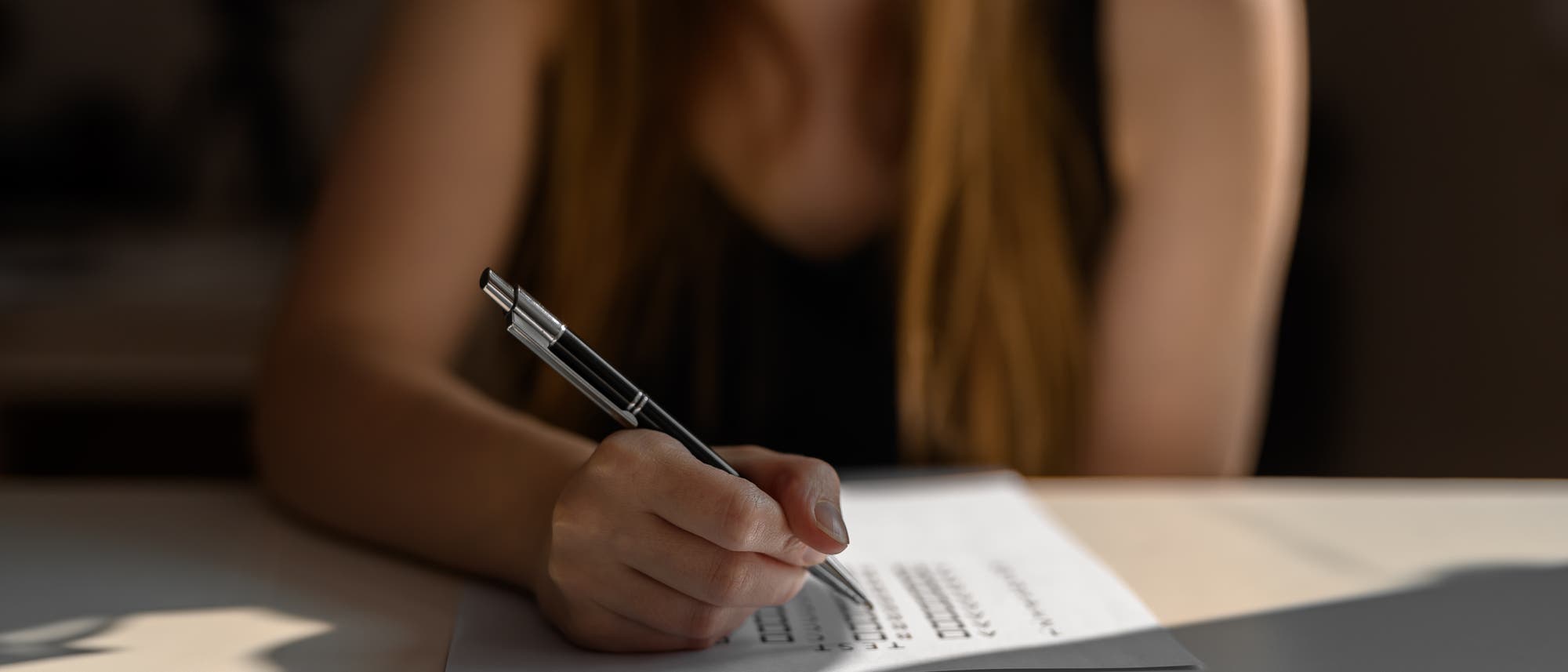 Eine Person sitzt an einem Tisch und schreibt mit einem Stift auf ein Blatt Papier. Das Bild ist in einem warmen Licht gehalten, das die Szene beleuchtet. Der Fokus liegt auf der Hand, die den Stift hält, während der Rest der Person unscharf im Hintergrund bleibt. Eine Person sitzt an einem Tisch und schreibt mit einem Stift auf ein Blatt Papier. Das Bild ist in einem warmen Licht gehalten, das die Szene beleuchtet. Der Fokus liegt auf der Hand, die den Stift hält, während der Rest der Person unscharf im Hintergrund bleibt.