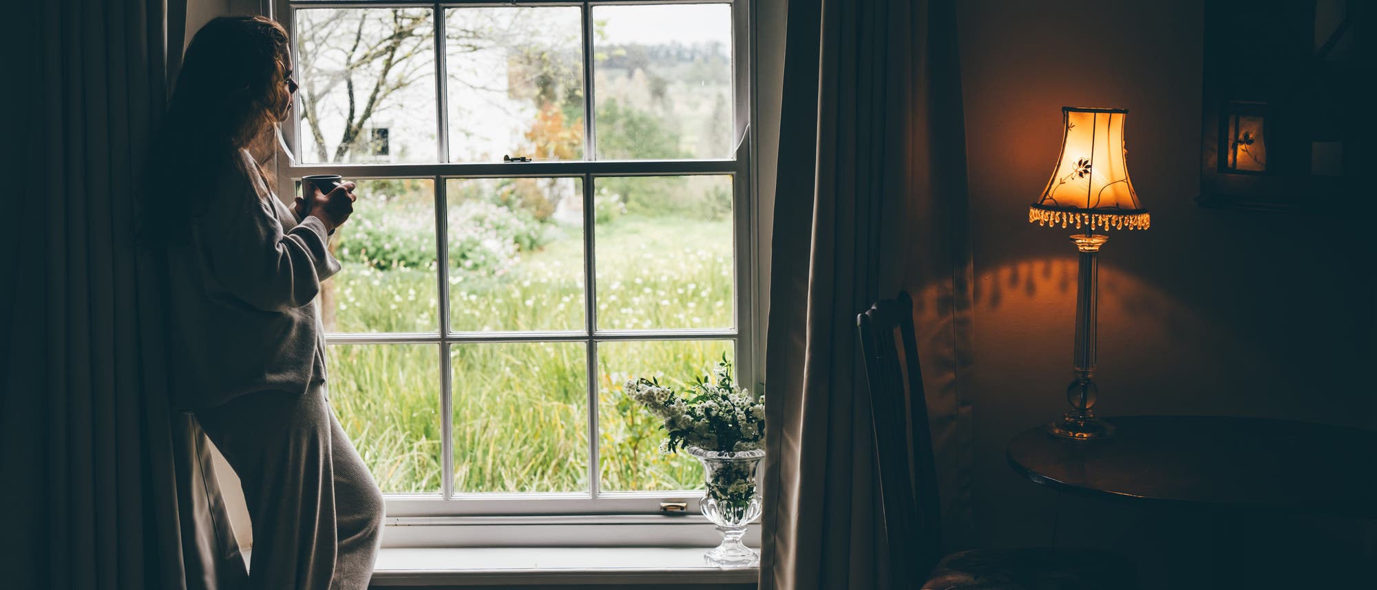 Eine Frau steht in einem gem&uuml;tlichen Raum und blickt aus einem gro&szlig;en Fenster auf eine gr&uuml;ne Landschaft. Sie h&auml;lt eine Tasse in der Hand. Neben dem Fenster spendet eine Tischlampe warmes Licht. Auf der Fensterbank steht eine Vase mit Blumen. Drau&szlig;en sind B&auml;ume und eine Wiese zu sehen.