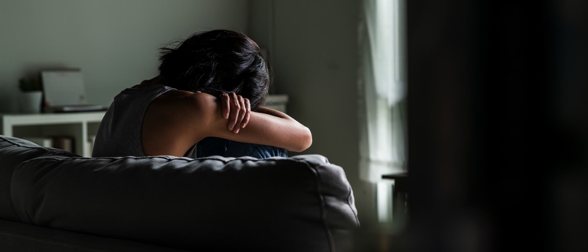 Eine Person sitzt auf einem Sofa in einem abgedunkelten Raum, den Kopf auf die Knie gelegt. Die Atmosphäre wirkt nachdenklich und melancholisch. Im Hintergrund sind ein Fenster mit leichtem Tageslicht und ein Regal zu sehen.