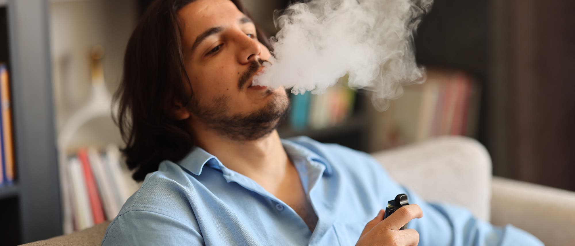 Eine Person sitzt entspannt auf einem Sofa und bl&auml;st eine Wolke aus Dampf aus, w&auml;hrend sie ein Vape-Ger&auml;t in der Hand h&auml;lt. Im Hintergrund sind B&uuml;cherregale zu sehen. Die Szene vermittelt eine entspannte Atmosph&auml;re in einem h&auml;uslichen Umfeld.