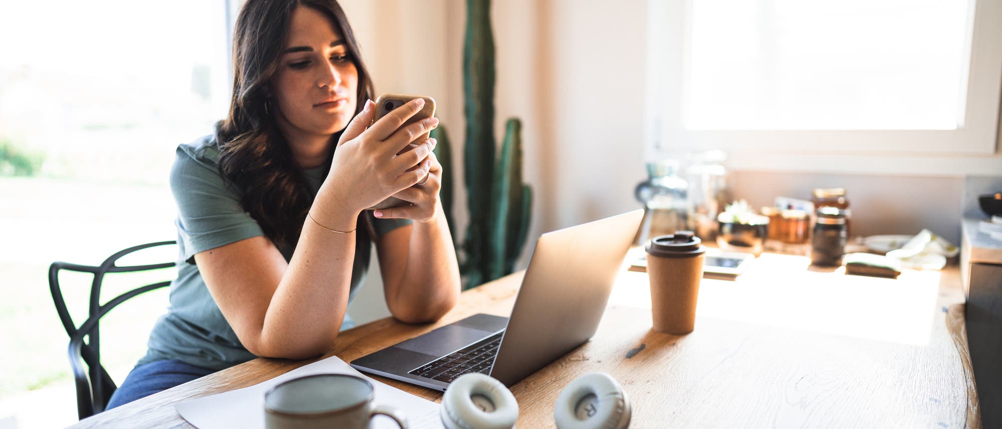 Eine Person sitzt an einem Holztisch und schaut auf ihr Smartphone. Auf dem Tisch befinden sich ein Laptop, eine Tasse, Kopfhörer und ein Kaffeebecher. Im Hintergrund steht ein Kaktus neben einem Fenster, durch das Tageslicht hereinfällt. Die Szene vermittelt eine entspannte Arbeits- oder Lernumgebung zu Hause.