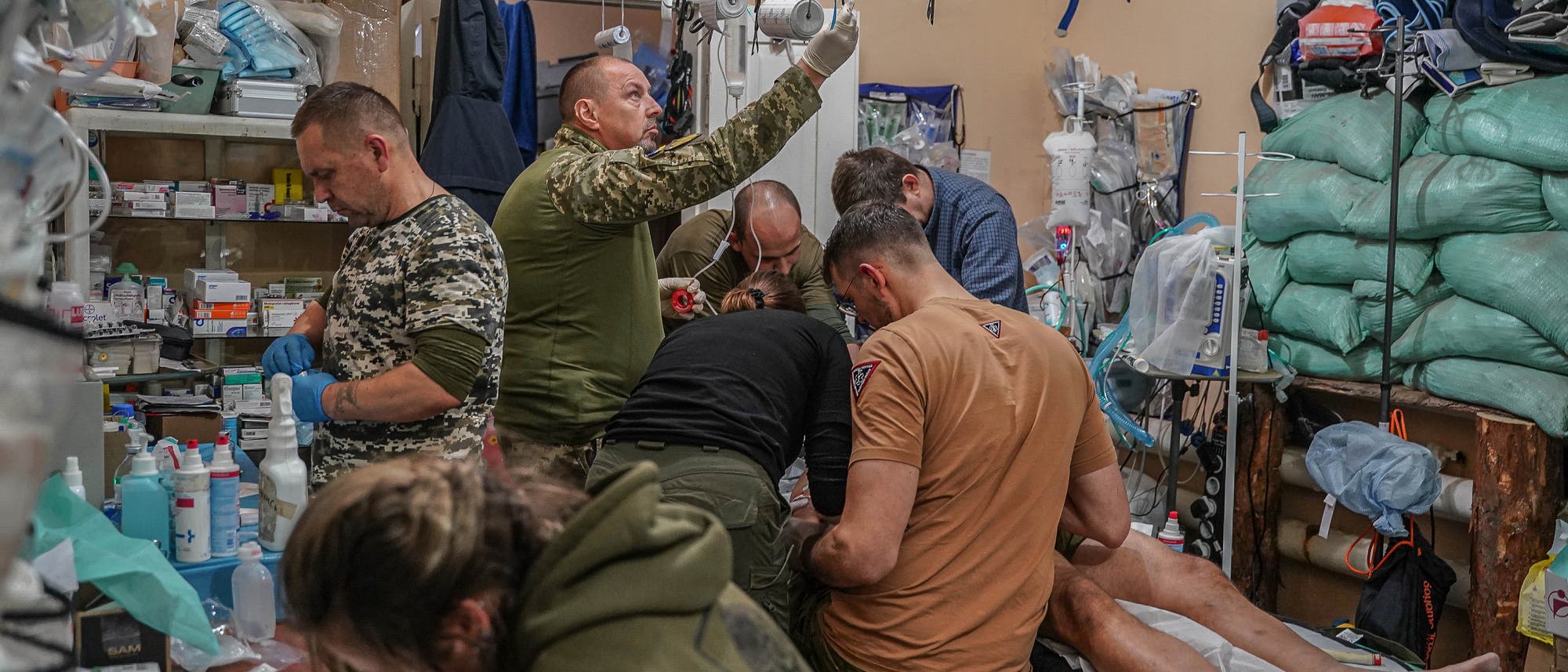 Eine Gruppe von Menschen in einem behelfsm&auml;&szlig;igen medizinischen Raum behandelt eine verletzte Person, die auf einer Liege liegt. Der Raum ist mit medizinischen Ger&auml;ten, Verb&auml;nden und Vorr&auml;ten gef&uuml;llt. Eine Person schreibt Notizen, w&auml;hrend andere sich um die medizinische Versorgung k&uuml;mmern. Die Szene vermittelt den Eindruck von Dringlichkeit und Teamarbeit in einer herausfordernden Umgebung.