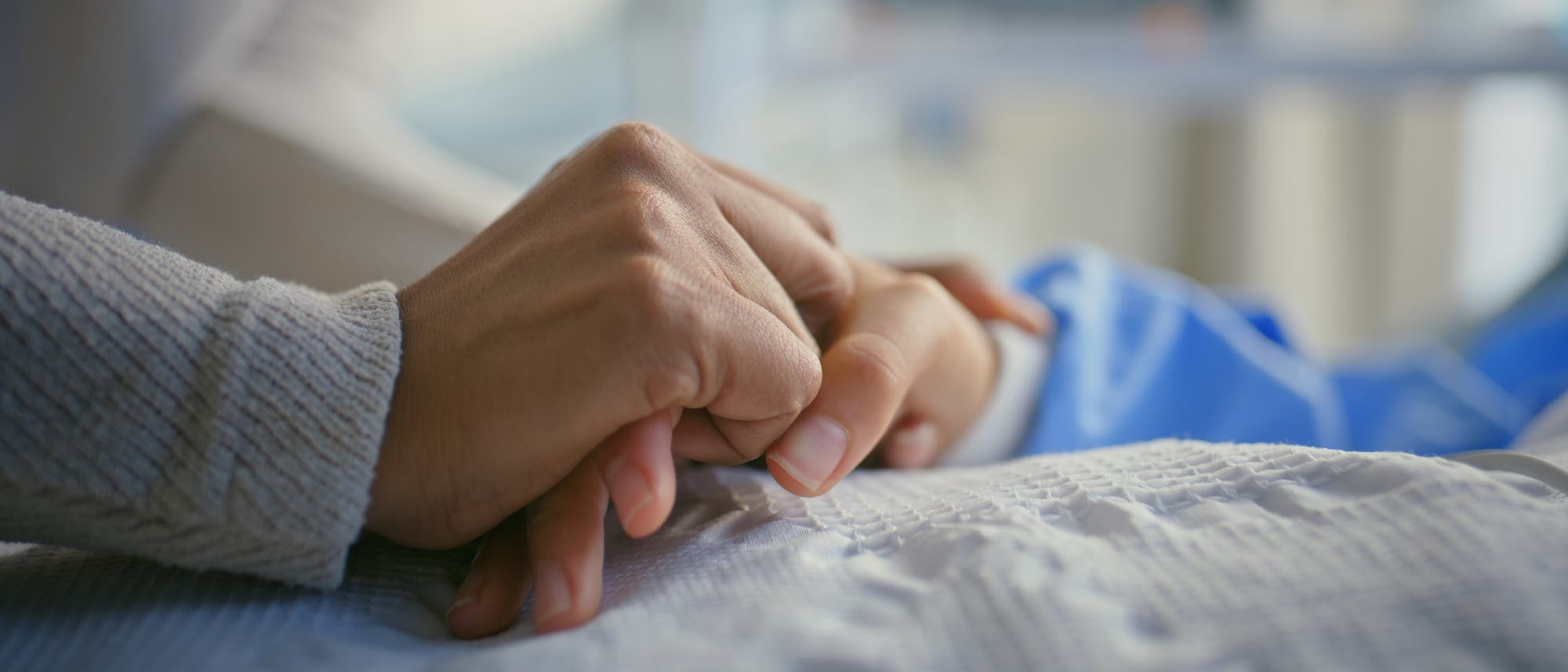 Eine Person hält die Hand einer anderen Person, die im Bett liegt. Im Hintergrund ist unscharf medizinisches Equipment zu erkennen. Die Szene vermittelt Fürsorge und Unterstützung in einer klinischen Umgebung.