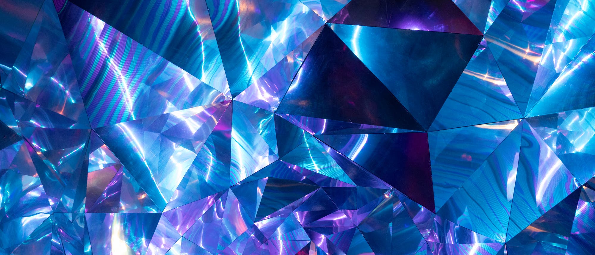 Ein abstraktes Kunstwerk mit einer Vielzahl von facettierten, prismatischen Formen in schimmernden Blau- und Violetttönen. Die geometrischen Muster reflektieren Licht und erzeugen einen kaleidoskopischen Effekt, der an Edelsteine oder Kristalle erinnert. Die Komposition vermittelt ein Gefühl von Tiefe und Bewegung.